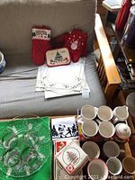 Photo shows Christmas napkins, oven mitts, cream and sugar set, mugs and decorative trays on table and couch.