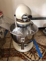 Side view showing cream-colored KitchenAid mixer with attached stainless steel mixing bowl and clear splash guard on tiled floor.