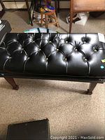 Full view of the double piano bench showing black tufted vinyl upholstery and wooden legs on a carpeted floor.