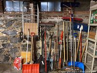 Full view showing all yard tools leaning against stone wall, including shovels, rakes, pitchfork, electric trimmer, and hand cart.