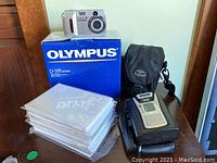 Overview of Olympus D-595 Zoom camera on box, Sony cassette recorder, camera case, and photo paper on table