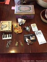 Top view photo of assorted cat-themed jewelry and figurines displayed on table with Lewis Sherry candy tin and other items.