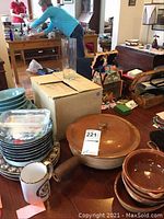 Overview of variety of kitchenware on table including stacks of blue ceramic plates, brown ceramic baking dishes with lids and handle, and a white mug