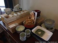 Full view of the lot showing tea cups, saucers, covered mugs in box, bowls, plates, and wooden utensils on table.