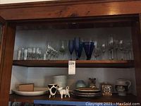Wide view of wood shelves showing various glassware and ceramics including clear and blue stemmed glasses, ceramic plates, bowls, and decorative ceramic pieces