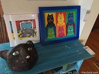 Photo showing the framed colorful cat print, paper mache cat mask on left, and two unframed cat prints in front.
