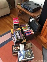 Panasonic VHS player placed on a small wooden table with VHS tapes stacked in and around a black carrying case on the floor.