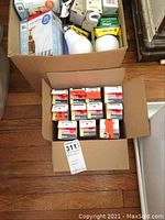 Close-up view of two open boxes showing diverse household light bulbs with multiple brands and wattage ratings, alongside cleaning products in bottles above the boxes.