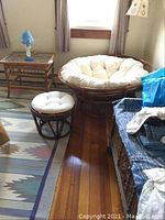 Papasan chair with cushion, matching ottoman, bamboo side table with glass top, and nearby blue glass lamp in room corner.