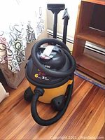 Side view of yellow and black Shop-Vac with coiled hose and nozzles attached, positioned on wooden floor near furniture.