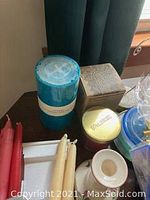 Shot of assorted candles stacked together including blue candle in packaging, boxed candle holder, gold tin candle, and paired taper candles.