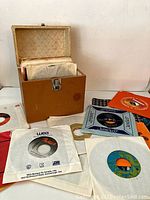 Photo of a vintage brown carrying case open, filled with 45 RPM vinyl records in paper sleeves, with some records spread out in front, showing a variety of label colors and designs.