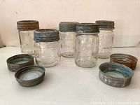 Five vintage clear glass Mason jars with embossed lettering and metal lids, plus four additional metal lids arranged in front.