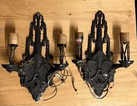 Pair of antique black metal wall sconces shown top down on wooden surface. Both sconces have two light holders each, with visible wiring and aged details.