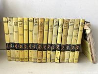 Photo showing 17 vintage Nancy Drew hardcover books standing in a row, titles visible on spines, with yellow dust jackets and wear.