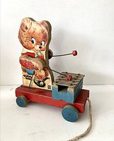 Front and side view of the Teddy Zillo pull toy showing the bear playing xylophone on red wooden base with blue wheels and pull string.