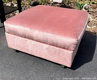 Full view of the dusty rose upholstered square ottoman with casters on the bottom, placed on black asphalt with background garden bed.