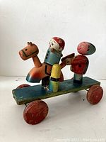 Side view of vintage wooden pull toy showing horse with rider and two standing figures on wheeled blue platform, signed and showing wear.