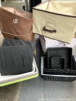 Various storage containers stacked together, including beige fabric bins with brown trim, black collapsible fabric boxes, and plastic baskets.