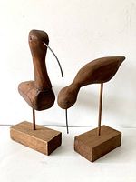 Pair of wood bird sculptures standing on rectangular wooden bases, each with curved metal beaks and small eyes.
