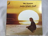 Album cover featuring a man sitting on the beach with a setting sun and a flying seagull graphic, titled Neil Diamond Jonathan Livingston Seagull.