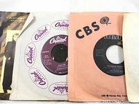 Photo showing multiple 45 RPM vinyl records with original sleeves from Capitol, CBS, and RCA labels.