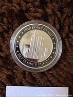 Front and raised image of Twin Towers with text 'United We Stand' and date 'September 11, 2001'. Coin placed on brown textured background with note card.