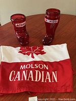 Two red glass Molson Canadian boot-shaped beer mugs and a Molson Canadian fabric banner laid out on a wooden surface.