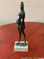 Bronze goddess sculpture front view on white marble base, showing slender female figure and artist's signature on base.