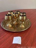 Top-down view of six brass goblets arranged on a round brass tray on a wooden surface, showing the polished brass finish and decorative tray edge.