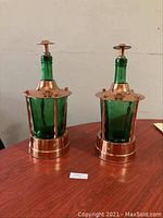Full view of two emerald green glass decanters encased in copper with wind-up music mechanisms visible at bases, both about 11 inches tall on a wooden surface.