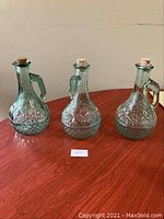 Three aquamarine glass bottles arranged on a wooden surface, each approximately 8 inches tall with raised design.