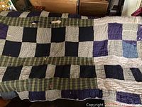 Photo showing the full spread of the quilt revealing patchwork squares in dark purple, black, beige, and green plaid fabric with visible signs of wear.