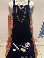Seven costume jewelry pieces displayed on a dark blue velvet fabric over a chair, including pearl-like necklaces, beaded necklaces, bracelets, and pins.
