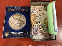 Blue hardcover H.G. Harris & Co. Explorer Worldwide Postage Stamp Album with vintage map design on cover beside open shoebox filled with loose postage stamps.