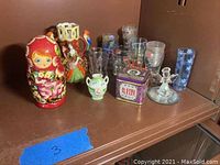 Wide view showing all items in the lot together on a shelf: Polish Matryoshka doll, Japanese Satsuma vase, shot glasses, angel figurine, small vases, and vintage tin box.