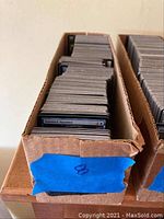 Two cardboard boxes filled with Magic: The Gathering cards, edges worn, visible blue tape on box end.