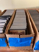 Three cardboard boxes filled with Magic: The Gathering cards, showing many cards stacked vertically, with one card label visible as 'Viashino Firstblade.'