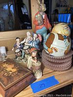 Photo showing Peter Rabbit figurine, American Revolution drummer and flag bearer figurines, wooden decorative box with floral inlay, and large ceramic cookie jar on top of stacked pink glass dishes.