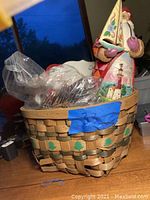 Photo showing woven basket with green tree accents filled with silverware in plastic bags and a Jim Shore Santa Claus figurine with lighthouse and floral details.