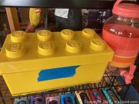 Yellow LEGO brick-shaped storage bin with eight studs on top and blue tape on the front, placed next to pink lemonade pitcher with a spout.