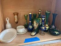 Shelf displaying multiple Japanese decorative glass vases in various colors with floral painted details and gold accents along with Lenox porcelain cups and saucers