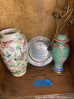 Photo shows ceramic vase with butterfly and flower design, Asian pottery lamp, and historic plate on wooden shelf.
