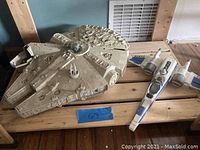 The set shows a beige Millennium Falcon toy model with detailed surface and an X-Wing fighter toy model with white and blue paint on a wooden surface.