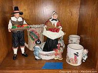 Two pilgrim figurines (male, female) holding harvest items (vegetables, apples), ceramic dish behind.