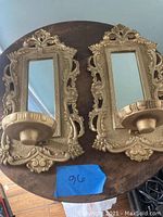 Pair of matching gold-toned wall fixtures with ornate design, rectangular mirrors, and attached bowl-shaped holders on a wooden circular table.