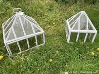 Two white garden housings, pyramid shaped with metal frames and glass panels, set on grass outdoors.