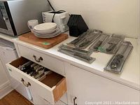 Overview of items on counter and inside open drawer showing cutlery block, packaged knives, toaster, breadboard, plates and bowls