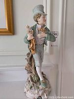 Full view of vintage male porcelain figurine standing on decorative base, dressed in period attire with a top hat and bouquet of flowers.