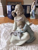 Glossy porcelain figurine showing two girls interacting and a cat resting on cloth at their feet
